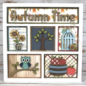 May include: A wooden framed shadow box with six compartments, each with a different autumn-themed design. The top compartment has the words "Autumn Time" in wooden letters. The other compartments feature a sunflower, a tree, a birdcage, an owl, a barrel of apples, and a single apple.