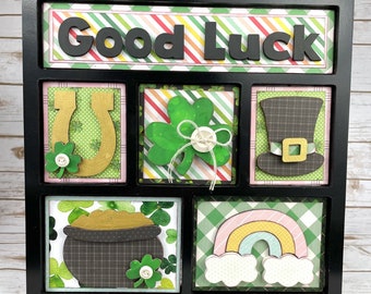 May include: A black shadow box with a white background and green shamrocks. The top of the box says "Good Luck". The box is divided into six sections with different images, including a horseshoe, a pot of gold, a rainbow, and a leprechaun hat.