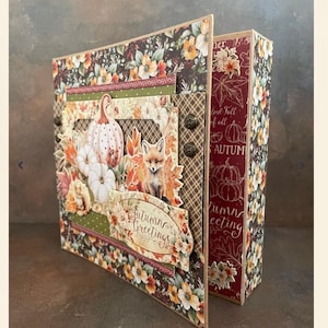 May include: A handmade scrapbook with a fall theme. The cover features a brown, green, and red floral pattern with a pumpkin, fox, and the text "Autumn Greetings".