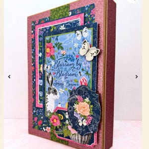 May include: A pink, blue, and green scrapbook paper with a floral design and a white rabbit. The paper has the text "Blossom by Blossom Spring Begins".