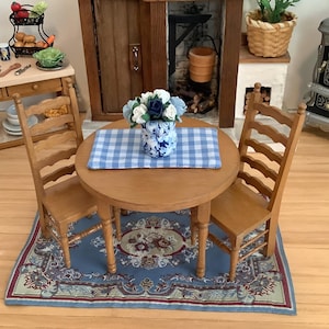 May include: A miniature wooden dining set featuring a round table and two ladder-back chairs. A blue and white checkered placemat sits on the table, topped with a blue and white floral vase containing white and dark blue fabric flowers.