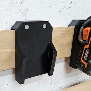 May include: A black plastic wall mount with two screws for hanging tools. The mount is designed to hold a power tool, such as a drill or screwdriver. The mount is attached to a wooden surface.