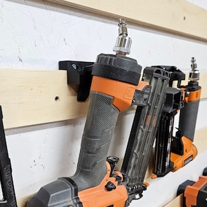 May include: Two orange and black nail guns with silver air hoses are mounted on a wooden wall. The nail guns are labeled "RIDGID".