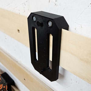May include: A black plastic wall mount with two screw holes and a slot for holding a tool. The mount is attached to a wooden wall.