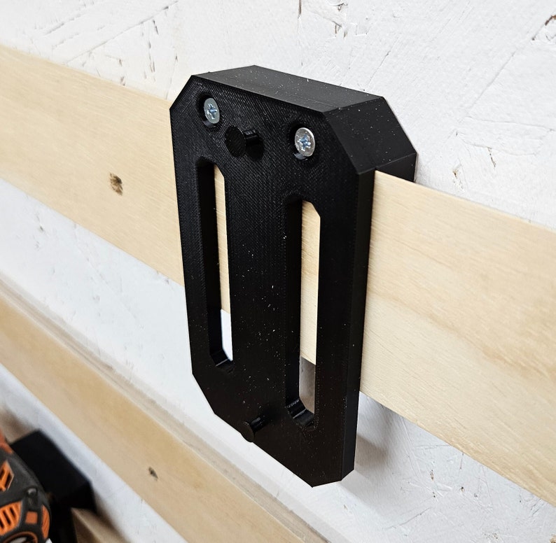 May include: A black plastic wall mount with two screw holes and a slot for holding a tool. The mount is attached to a wooden wall.