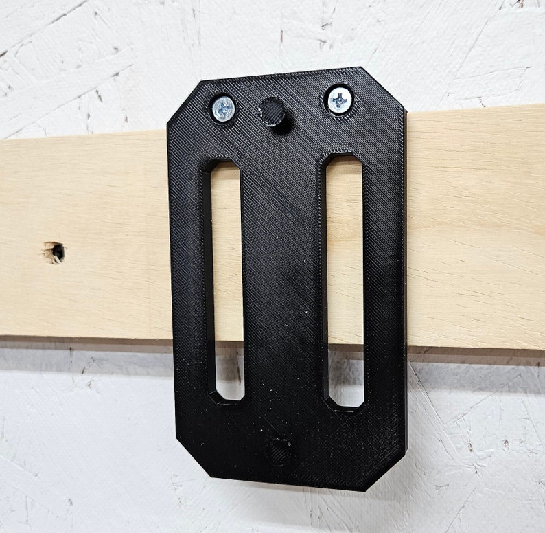 May include: Black plastic wall mount with two slots and three screw holes. The mount is attached to a light wood surface.