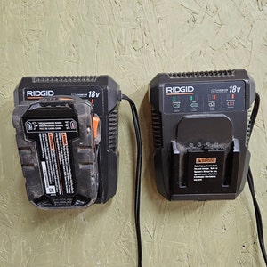 May include: Two black 18V Ridgid battery chargers with a black cord attached to each. The charger on the left has a battery pack attached to it. The charger on the right has a warning label on it.
