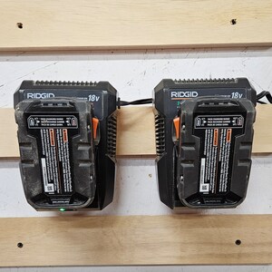 May include: Two black and orange RIDGID 18v battery chargers mounted on a wooden wall. The chargers have a label with the text "PACK CHARGING RANGE" and "PACK DE CARGA GAMA".