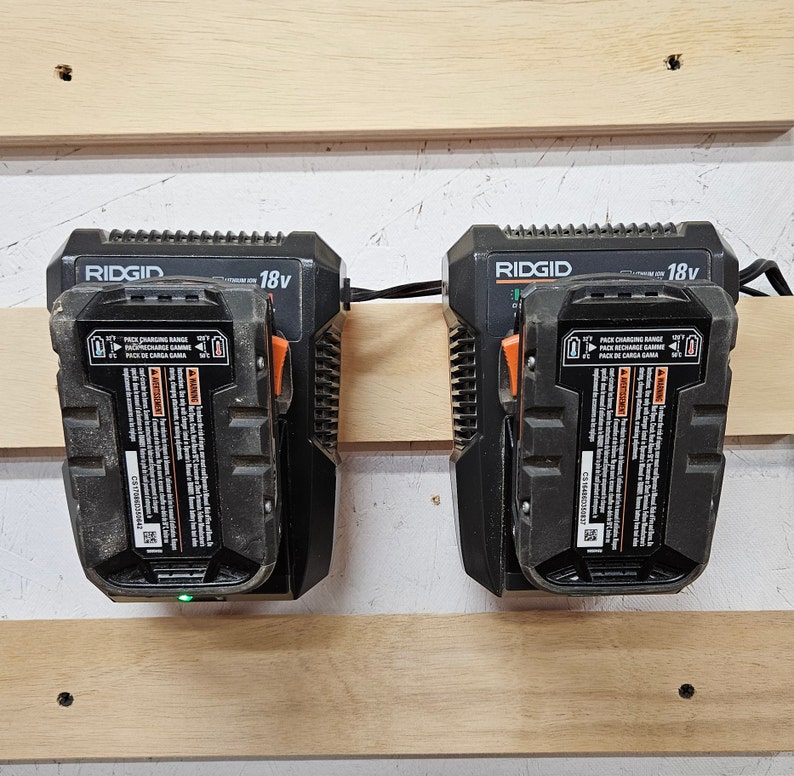 May include: Two black and orange RIDGID 18v battery chargers mounted on a wooden wall. The chargers have a label with the text "PACK CHARGING RANGE" and "PACK DE CARGA GAMA".