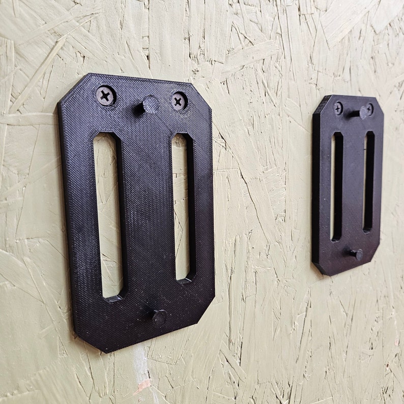 May include: Two black plastic wall mounts with slots for holding items. The mounts are attached to a light beige wall.