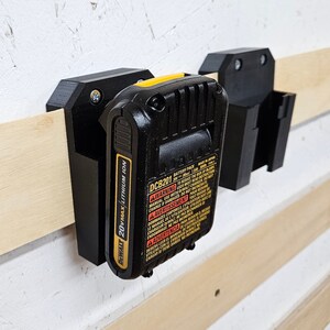 May include: A black and yellow DeWalt 20VMAX lithium-ion battery pack holder mounted on a wall. The holder has a warning label that reads "WARNING: Battery Pack. Made in Japan. To reduce the risk of injury, read the instruction manual. Use with DeWalt charger only. Do not charge if damaged. Do not incinerate."