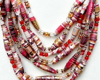 Puede incluir: Collar de varias hebras con cuentas de papel en tonos rojos, rosas y dorados. Las cuentas son cilíndricas y están ensartadas con cuentas rojas y doradas más pequeñas. El collar tiene un cordón de fibra natural.
