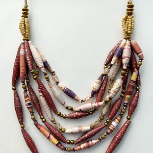 May include: A multi-strand necklace made of rolled paper beads in shades of brown, cream, and purple. The beads are interspersed with gold and clear beads. The necklace is a statement piece and would be a great addition to any wardrobe.