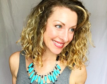 May include: A woman with curly blonde hair is wearing a turquoise and black beaded necklace. The necklace has a cascading design with long, thin beads.