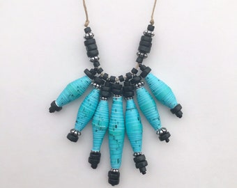 May include: A turquoise and black beaded necklace with a long, cascading design. The necklace features a series of turquoise paper beads and black wooden beads.