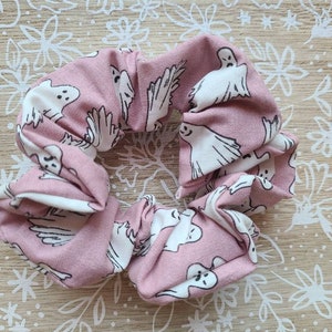 Op de afbeelding: Een roze scrunchie met een herhalend patroon van witte spookillustraties. De stof is geplooid en gerimpeld, waardoor een textuur ontstaat. De scrunchie is ontworpen voor haarstyling.