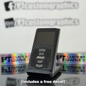 May include: A black air suspension control unit with a carbon fiber pattern and a digital display. The unit is sitting on a black charging stand. The text "AIRLIFT PERFORMANCE" is printed on the unit. The text "[includes a free decal]" is printed below the unit.