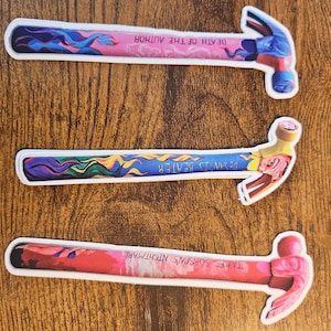 May include: Three hammer stickers with colorful designs. The top sticker has a pink handle with the text "DEATH OF THE AUTHOR". The middle sticker has a blue handle with the text "DESANTIS BEATER". The bottom sticker has a red handle with the text "JAMES SOBSON'S NIGHTMARE".