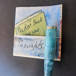 May include: A turquoise and gold fountain pen next to a watercolor illustration of a pocket book. The illustration has the words "Pocket Book for your Thoughts" written on it. The pen has a marbled design.