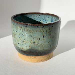 Könnte beinhalten: Ein Keramikbecher mit einer gesprenkelten türkis-blauen Glasur, die in einen dunkelbraunen Boden übergeht. Der Rand ist dunkelbraun und der untere Teil besteht aus unglasierter, texturierter beiger Tonerde. Das Innere hat ein glattes, dunkelblaues Finish.