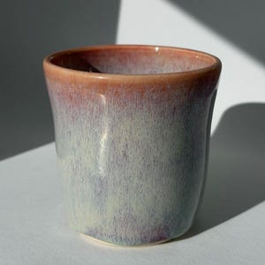 Könnte beinhalten: Ein Keramikbecher mit einer glatten, glänzenden Glasur, die von einem warmen, rötlich-orangen Farbton am Rand zu einer weichen, gesprenkelten Mischung aus blassblau, lavendel und cremefarbenen Tönen am zylindrischen Körper mit leicht ausgestellter Öffnung übergeht.