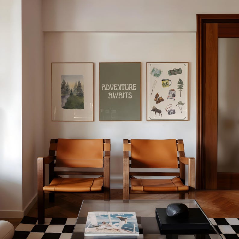 May include: Three framed prints on a white wall, two with nature scenes and one with the text "Adventure Awaits". Two brown leather chairs with wooden frames are in front of the wall, and a glass coffee table with a book and a black stone object is in front of the chairs.