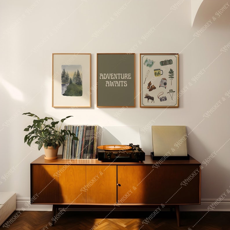 May include: Three framed prints on a white wall above a wooden dresser. The prints are all vertical and feature nature-themed designs. The leftmost print shows a watercolor illustration of a forest scene. The middle print has a green background with the text "Adventure Awaits" in white. The rightmost print features a collection of camping and hiking gear, including a compass, a backpack, a mug, and a pair of boots.