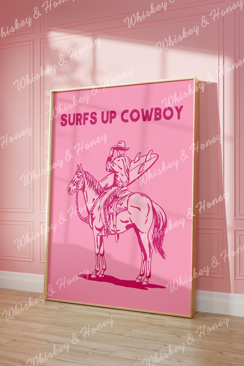 May include: A pink poster featuring a cowboy riding a horse and holding a surfboard. The text "SURFS UP COWBOY" is printed in pink on the poster.