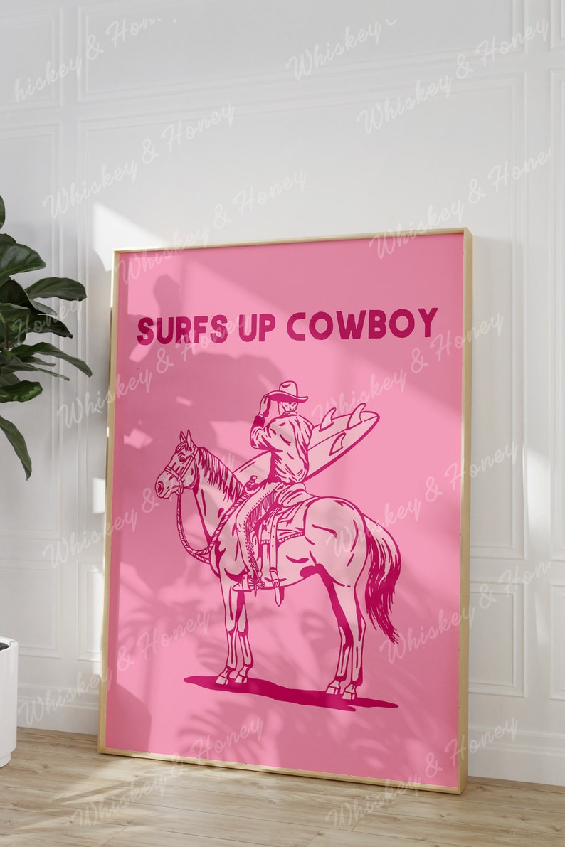 May include: A pink framed poster with the text "SURFS UP COWBOY" in bold pink letters. A pink illustration of a cowboy riding a horse while holding a surfboard.