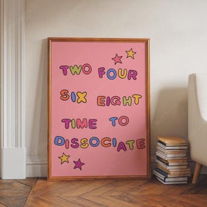 Puede incluir: Un cuadro rosa enmarcado con las palabras "TWO FOUR SIX EIGHT TIME TO DISSOCIATE" en letras de burbujas de colores. La impresión incluye ilustraciones de estrellas y se exhibe en una habitación con una pila de libros.