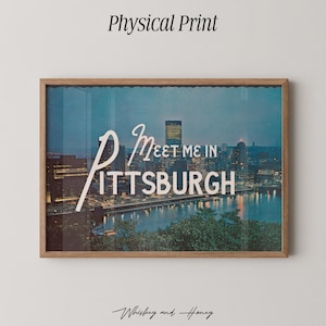 Op de afbeelding: Een ingelijste print met een nachtelijk stadsbeeld van Pittsburgh, Pennsylvania, met de woorden "Meet Me in Pittsburgh" in witte letters. De print zit in een houten lijst en bevat bovenaan de tekst "Physical Print".