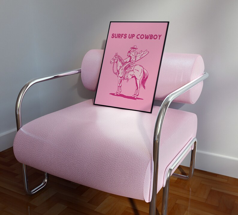 May include: A pink framed print with the text "Surfs Up Cowboy" in white letters. The print features a line drawing of a cowboy riding a horse while holding a surfboard.