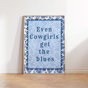 May include: A framed print with a blue and white floral border. The text inside the frame reads "Even Cowgirls get the blues".