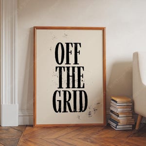 May include: A framed print with the words "OFF THE GRID" in bold black letters. The print has a light beige background and a brown wooden frame. The print is leaning against a wall on a wooden floor.