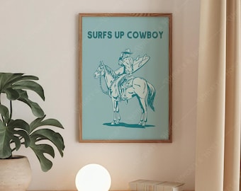May include: A framed poster with the text "SURFS UP COWBOY" above a turquoise illustration of a cowboy riding a horse while holding a surfboard. The poster is in a wooden frame and hangs on a light-colored wall.