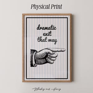 May include: Framed print featuring a black and white illustration of a hand pointing right, with the words "dramatic exit that way." The print is in a light wood frame and has a striped background.
