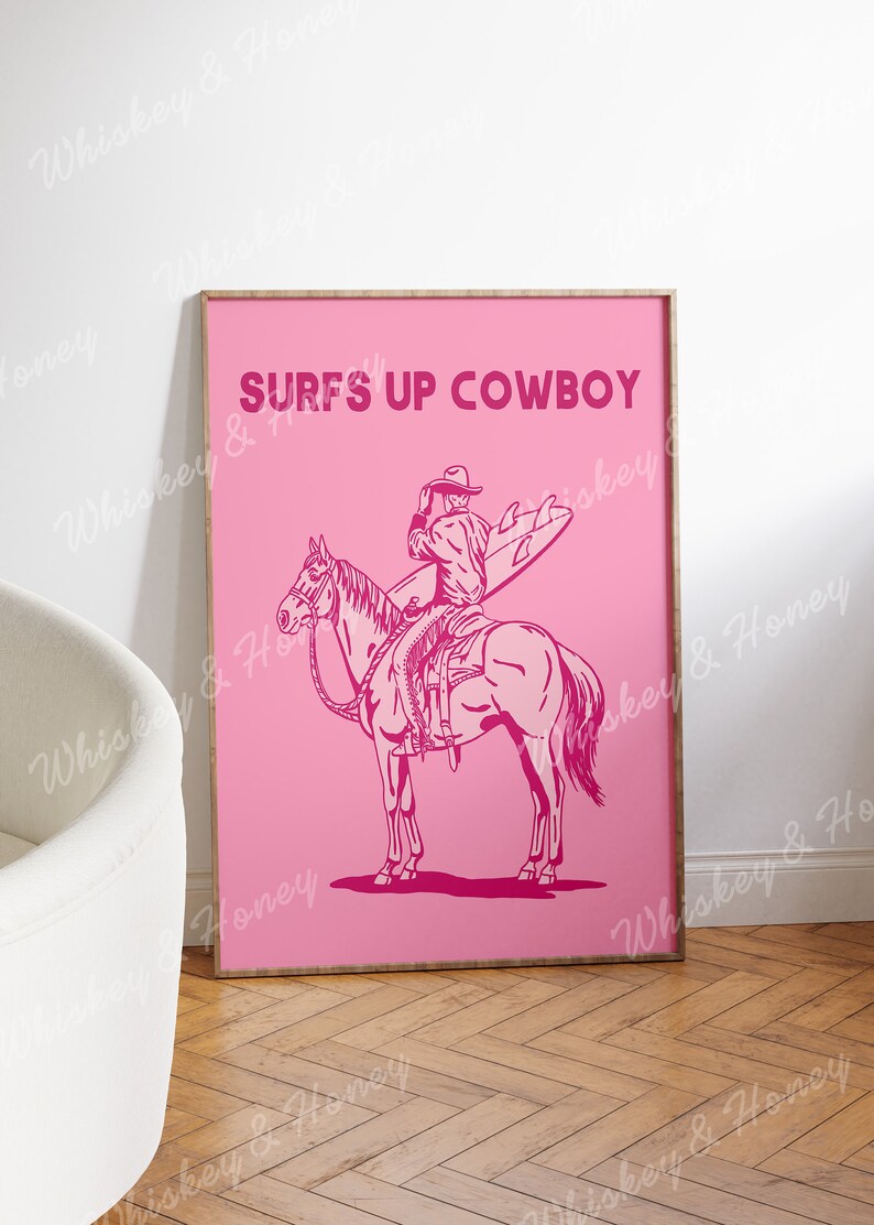 May include: A pink poster featuring a cowboy riding a horse and holding a surfboard. The text "SURFS UP COWBOY" is printed in bold pink letters.