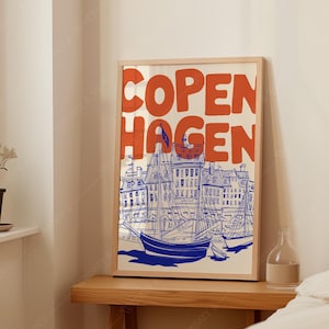 Pode incluir: Uma impressão artística emoldurada com a palavra "COPENHAGEN" em laranja. A impressão mostra uma ilustração azul e branca de uma cena portuária com barcos e edifícios. A obra de arte é exibida numa mesa de madeira.