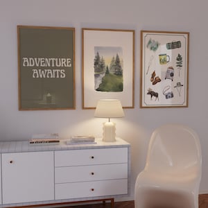 May include: Three framed prints on a white wall, a white chair, and a white dresser with three drawers. The prints are all different, but they all have a nature theme. The first print says "Adventure Awaits" in a retro font. The second print is a watercolor painting of a forest scene. The third print is a collection of camping-themed illustrations.