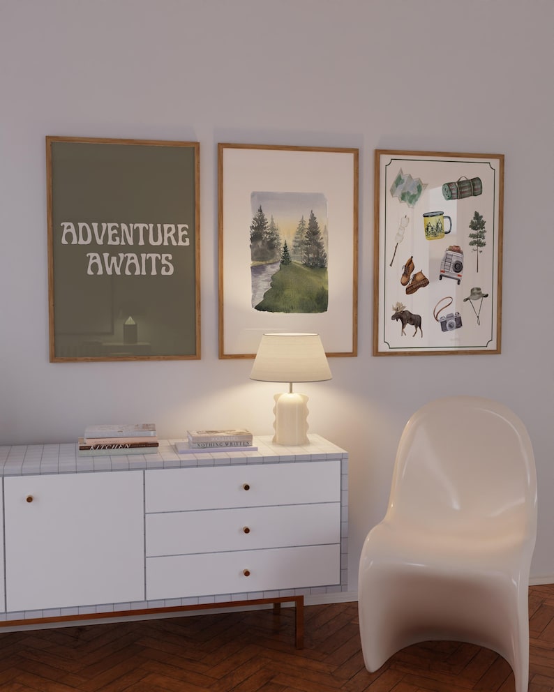 May include: Three framed prints on a white wall, a white chair, and a white dresser with three drawers. The prints are all different, but they all have a nature theme. The first print says "Adventure Awaits" in a retro font. The second print is a watercolor painting of a forest scene. The third print is a collection of camping-themed illustrations.