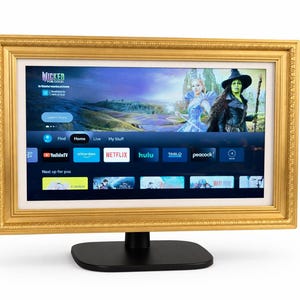 May include: A gold-framed television displaying a streaming service interface. The screen shows a scene from "Wicked" and various app icons. The TV is mounted on a black stand, creating a decorative and functional piece.