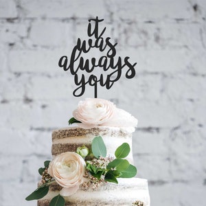 May include: A three-tiered cake with a semi-naked frosting design, adorned with light pink flowers and greenery. A black cake topper reads "it was always you". The cake is set against a white brick wall background.