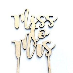 May include: A wooden cake topper with the words "Miss to Mrs" in a cursive font. The topper is made of light-colored wood and has two stakes for insertion into a cake. The design is suitable for a wedding or bridal shower.