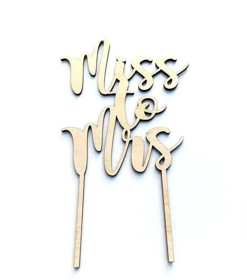 May include: A wooden cake topper with the words "Miss to Mrs" in a cursive font. The topper is made of light-colored wood and has two stakes for insertion into a cake. The design is suitable for a wedding or bridal shower.