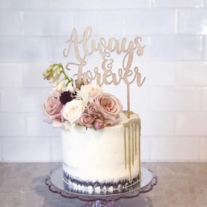 May include: A round white cake with a floral arrangement of pink and white roses, and a wooden topper that reads "Always & Forever." The cake has gold icing drips and sits on a clear glass cake stand.