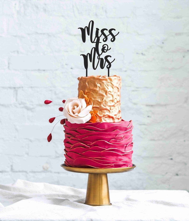 May include: A two-tiered wedding cake with a gold top tier and a red bottom tier. The cake is decorated with a white rose and a black cake topper that reads "Miss to Mrs". The cake sits on a gold cake stand.