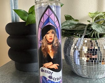 Könnte beinhalten: Eine Glaskerze mit einem violetten, blauen und rosafarbenen Buntglasdesign. Die Kerze zeigt ein Porträt von Stevie Nicks mit dem Text "Saint Stevie" unten.