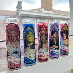 Puede incluir: Cinco tarros de velas de vidrio con dise&ntilde;os de vidrieras de colores e im&aacute;genes de Dolly Parton, Shrek, Taylor Swift, Stevie Nicks y Chappelle. Cada tarro tiene una etiqueta que dice "Saint" seguido del nombre de la persona representada.