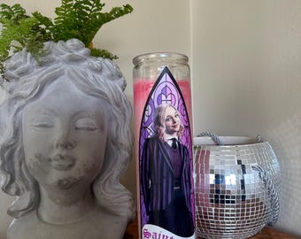 Könnte beinhalten: Eine Glaskerze mit rosa Innenraum und einem Etikett mit einem Porträt einer Person in einem schwarz-lila gestreiften Blazer. Auf dem Etikett steht "Saint Enid" in gotischer Schrift. Eine graue Statue eines Gesichts und ein Discokugel-Topf sind ebenfalls sichtbar.