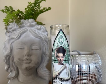 Könnte beinhalten: Eine weiße Kerze mit einem Saint Frankenstein-Etikett mit einem Porträt der Braut von Frankenstein. Ein grauer Steinkopf-Pflanztopf mit Grün und ein silberner Discokugel-Kerzenhalter sind ebenfalls zu sehen.